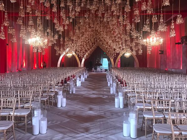 An ornate indoor venue set up for an event with rows of white chairs, hanging chandeliers, and glowing arches, creating an elegant and festive ambiance.