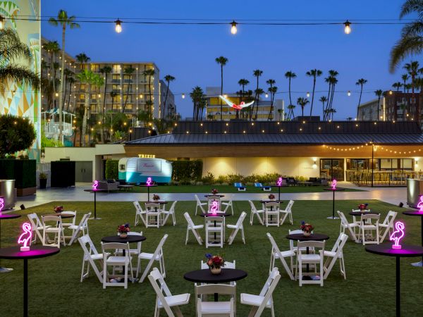 An outdoor venue with string lights, tables, chairs, neon signs, and palm trees at dusk.