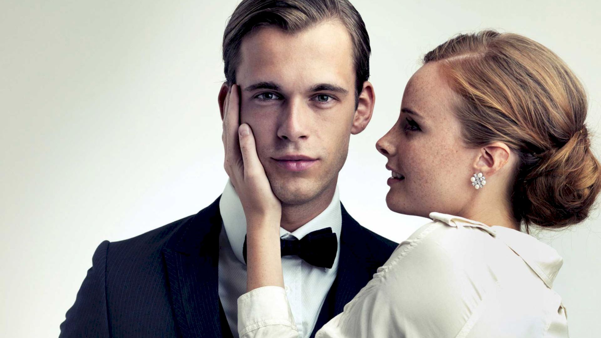 A woman, dressed elegantly, holds a man's face tenderly while he looks straight ahead wearing a suit and bow tie.