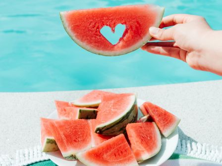 A hand holds a watermelon slice with a heart-shaped cutout over a pool; a plate of watermelon chunks rests on a colorful towel by the pool.