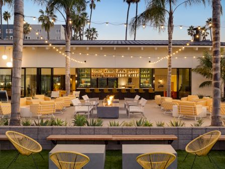 This image shows an outdoor patio with modern seating, string lights, palm trees, and a central fire pit, all facing a bar with stools.