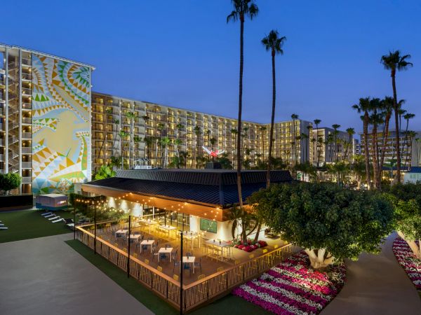 The image shows an outdoor dining area with lights, surrounded by trees and flowers, next to a tall hotel building with a mural and palm trees.