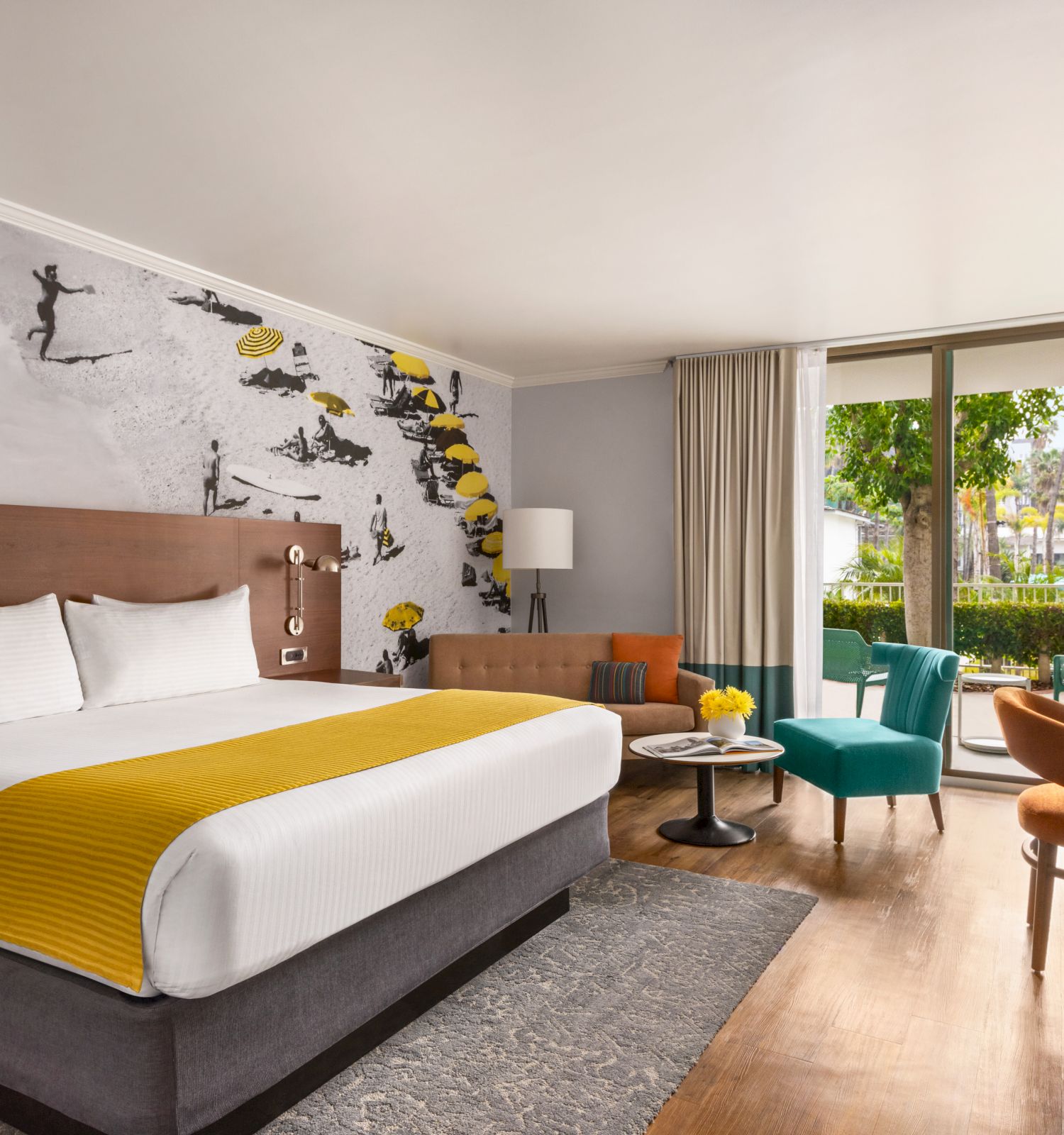 A modern hotel room with a king-size bed, yellow accents, a sofa, armchairs, a wall mural, a TV, and a large window overlooking greenery in the Best Resort in San Diego with a waterslide.