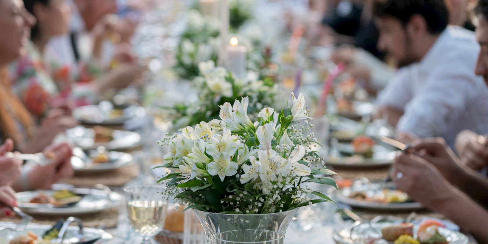 People are dining at a long table adorned with white flowers, enjoying a meal together in a communal setting outdoors at a beautiful and unique wedding venue in San Diego.