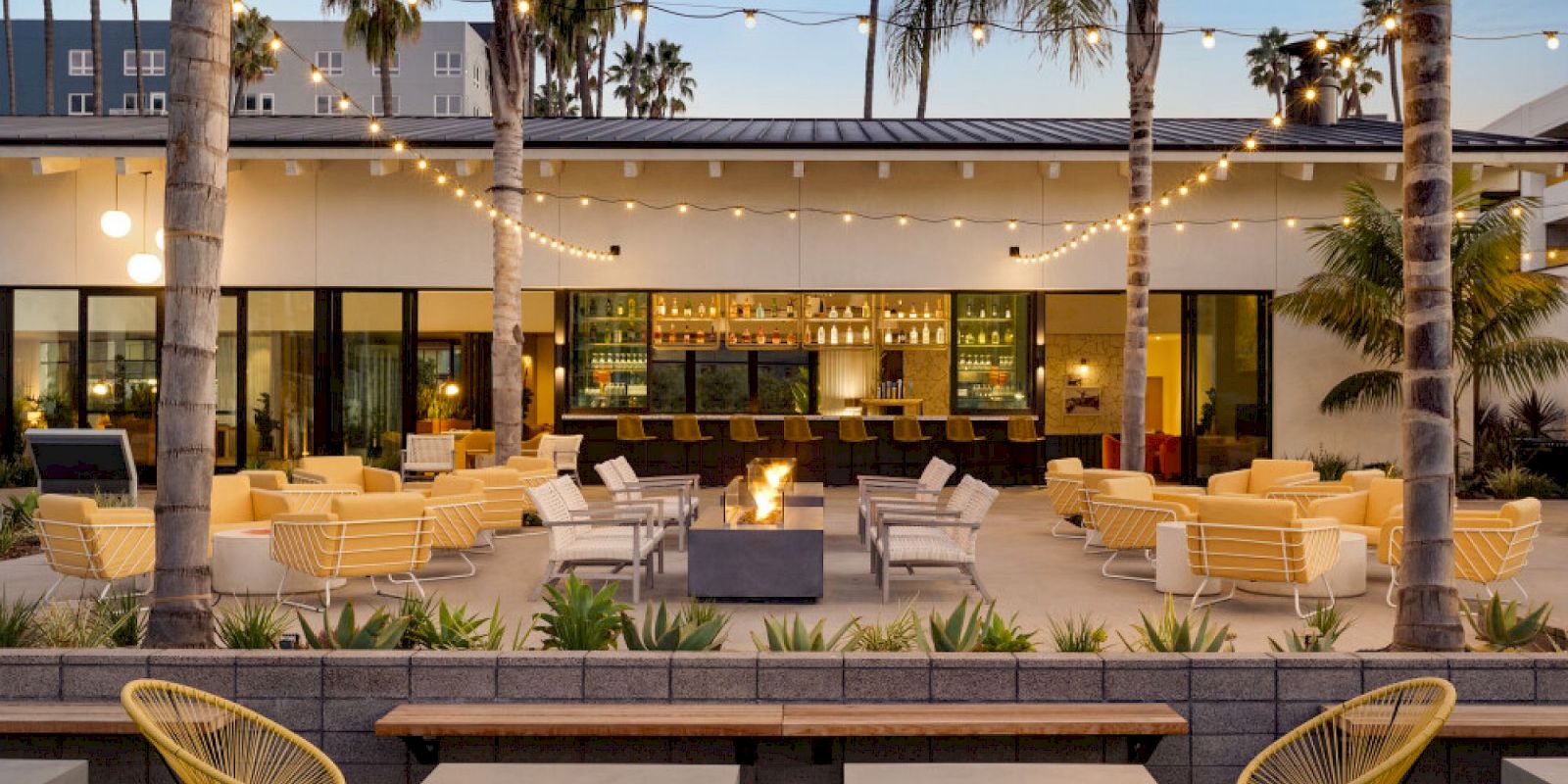 The image shows an outdoor patio with modern seating, string lights, palm trees, a central fire pit, and a bar area in the background at the best bar in Mission Valley and San Diego