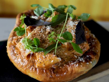 The image shows a gourmet pizza topped with greens, cheese, and other ingredients, served on a wooden board and black cloth napkin.