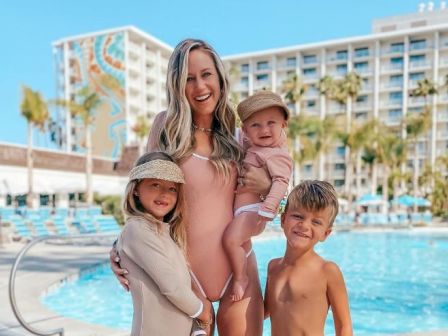 A woman and three children smile poolside, with a hotel building and palm trees in the background. All are in swimwear and looking happy.