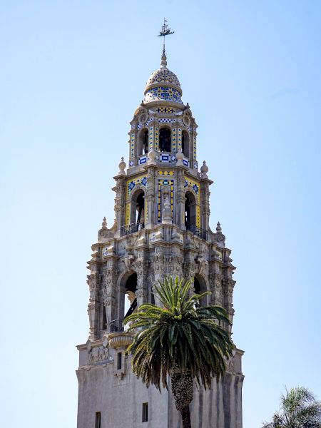 A tall, ornate tower with intricate architectural details and a palm tree in the foreground against a clear blue sky.
