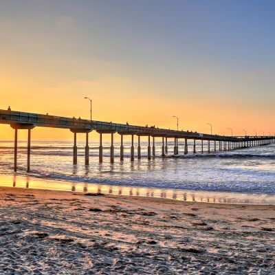 A sunny sunset at the beach with a long pier over the waves and a surfer holding a board near the shore.