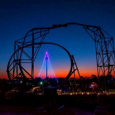 A massive roller coaster ride glows with purple and pink lights against a sunset sky over dark grounds at SeaWorld San Diego.