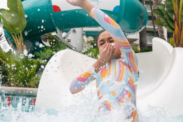 A person in colorful swimwear emerges from a water slide into a splash pool, covering their mouth with water splashing around them.
