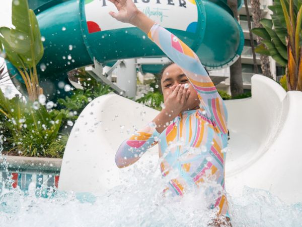 A person in colorful swimwear emerges from a water slide into a splash pool, covering their mouth with water splashing around them.