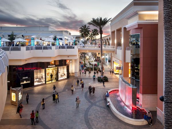 A lively outdoor shopping mall at dusk, with palm trees, bright storefronts like Prada, and shoppers walking past a glowing fountain.