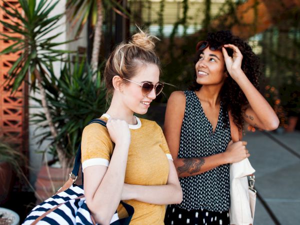 Two people are outdoors, smiling and looking happy. One is wearing sunglasses and carrying a striped bag; the other is holding a white bag.