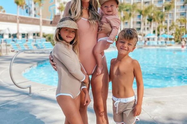 A woman and three children are posing by a swimming pool in swimsuits, with a hotel building and some palm trees in the background.