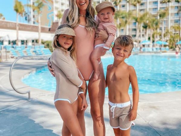 A woman and three children are posing by a swimming pool in swimsuits, with a hotel building and some palm trees in the background.
