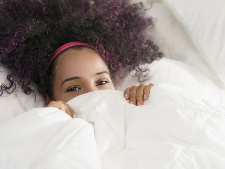 A person with curly hair is peeking out from under a white blanket, showing a small part of their face while appearing cozy in bed.
