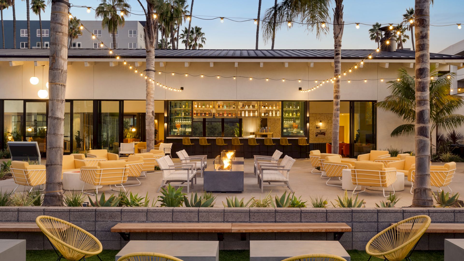 Outdoor lounge area with string lights, fire pit, yellow and white seating, and a bar in the background, surrounded by palm trees.