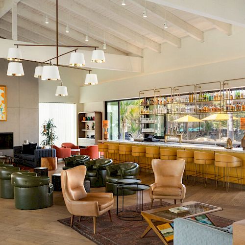A modern, stylish lounge featuring leather chairs, green stools, a bar with yellow stools, art, large windows, and a fireplace with a cozy ambiance.