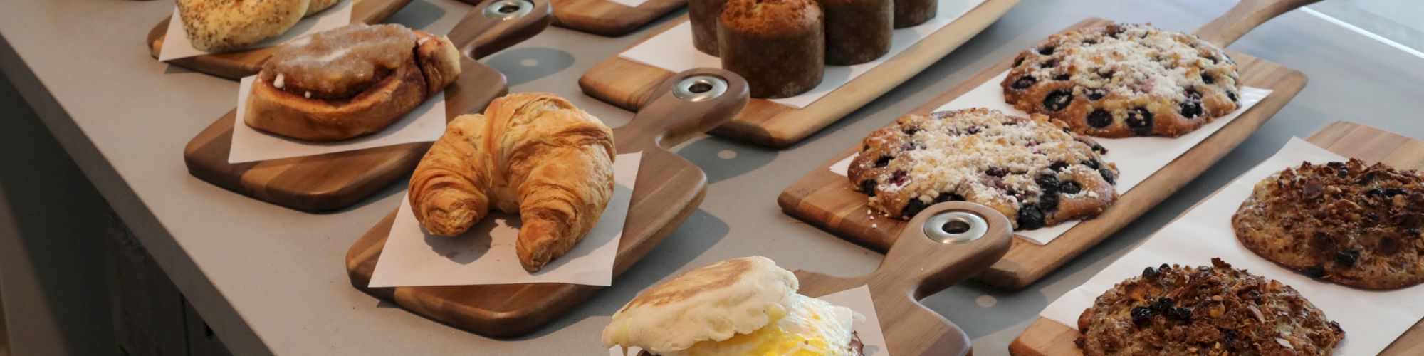 A variety of baked goods displayed on wooden boards, including pastries, croissants, muffins, cookies, and sandwiches, in a well-lit area.
