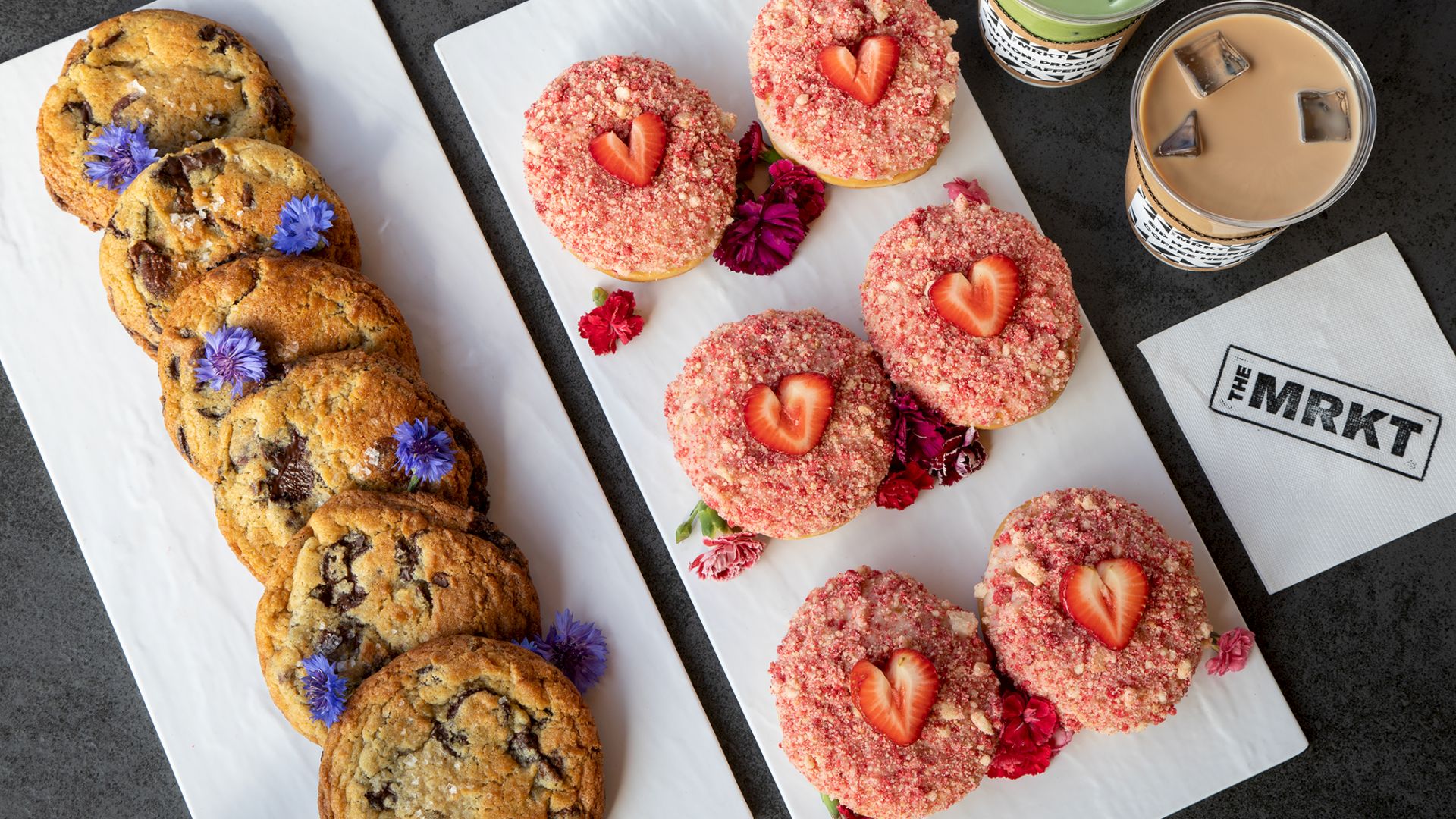 A table of iced strawberry doughnuts and chocolate-chip cookies beside two cups of coffee and a chalk-marked napkin.
