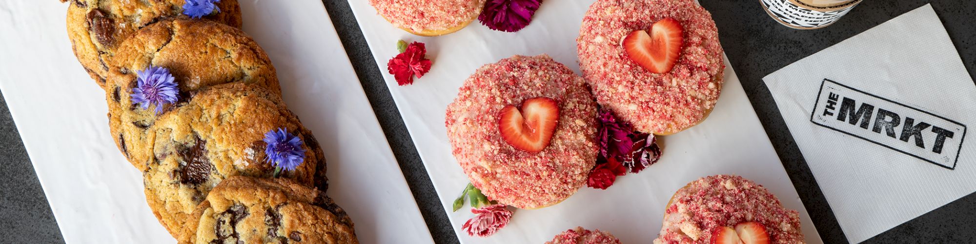 A table of iced strawberry doughnuts and chocolate-chip cookies beside two cups of coffee and a chalk-marked napkin.