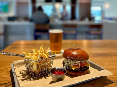 A burger and fries with a beer sitting on a table in a restaurant, with a blurred background showing people and a TV screen.