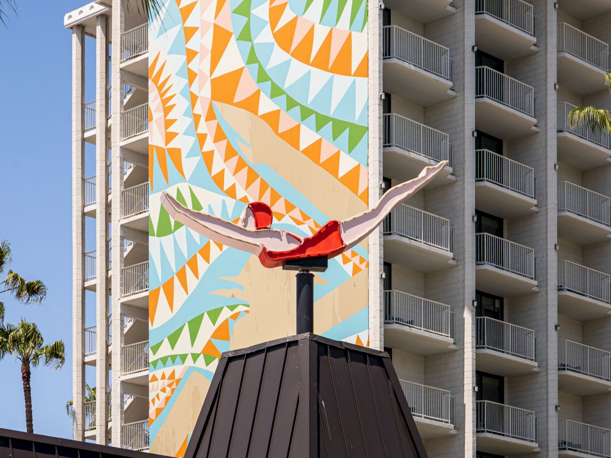 A hotel building features colorful geometric patterns on its facade and a roof sculpture of a stylized bird. Palm trees are also visible.