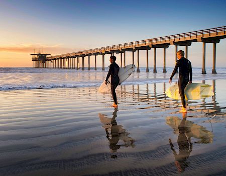Two surfers walk along a reflective beach at sunset, carrying their surfboards near a long pier extending into calm waves. Ends with a period.