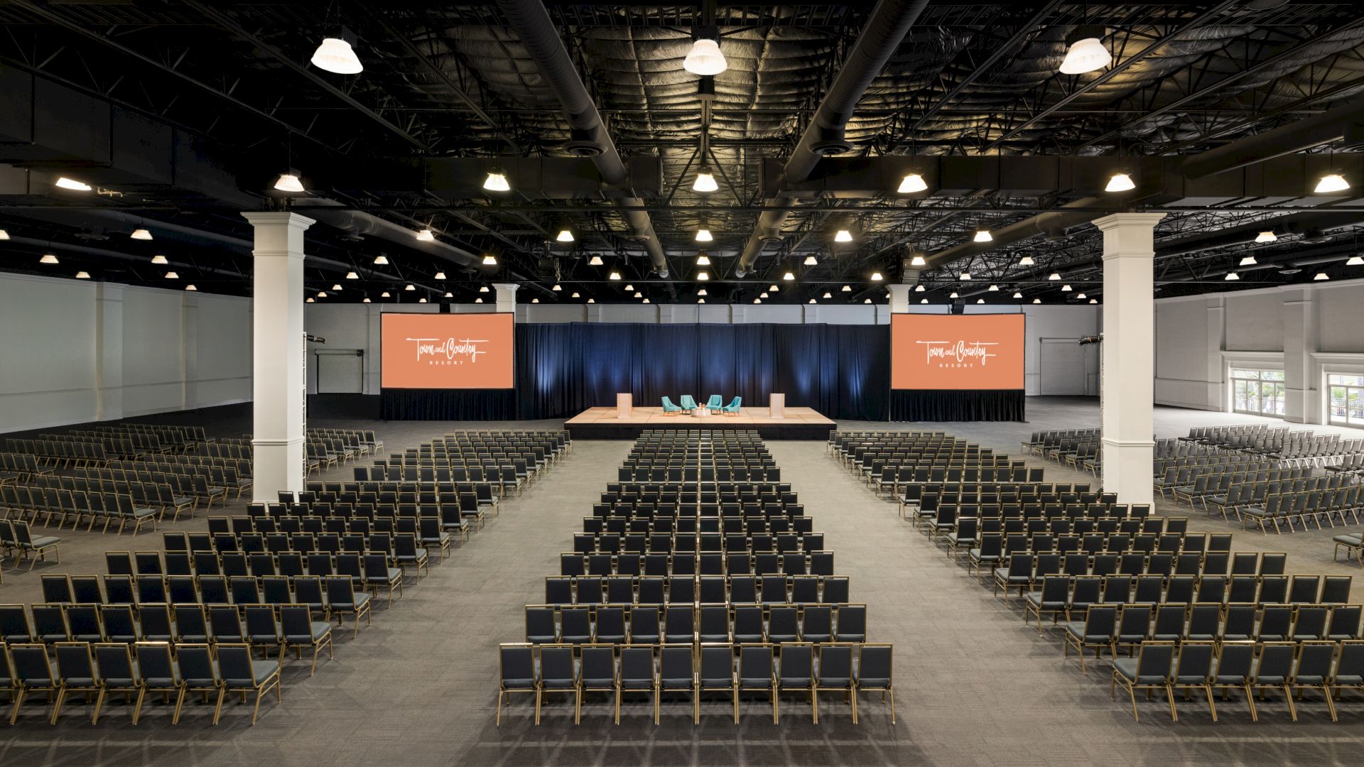 The image shows a large, empty conference room with rows of chairs facing a stage with a podium and screens on each side, illuminated by ceiling lights.