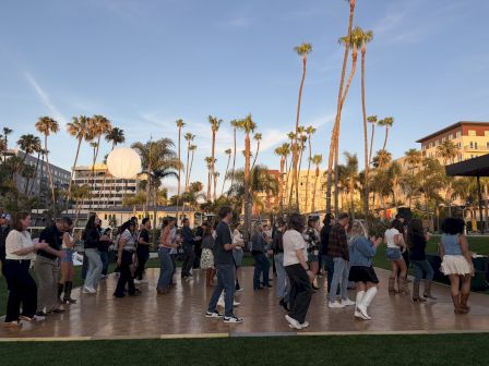 People lined up in a sunny palm-lined plaza, casually walking, chatting, and posing near a fountain and buildings.