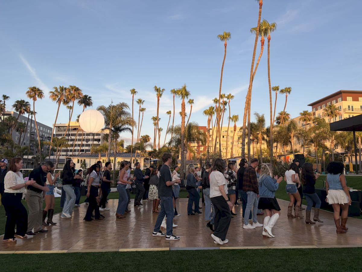 People gather outdoors on a sunny campus lined with tall palm trees and warm-colored buildings, chatting and walking by.