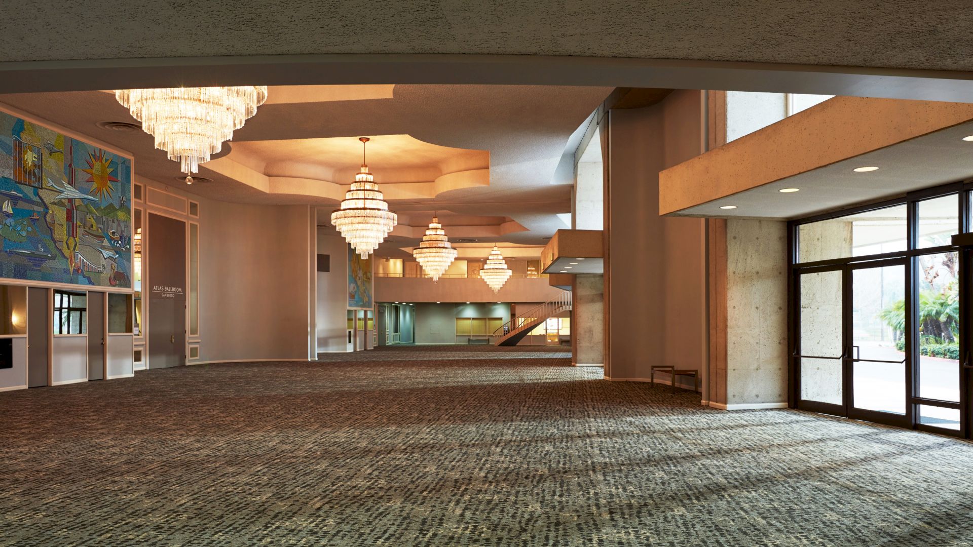 A spacious, well-lit interior with carpet flooring, large chandeliers, wall art, extensive ceiling architecture, and a glass door entrance.