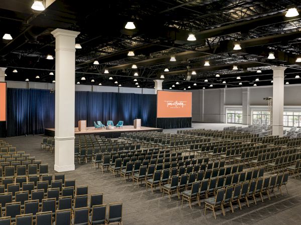 A large conference room with rows of chairs, a stage with chairs, and two large screens displaying text on either side of the stage.