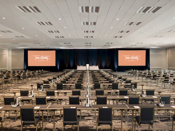 A large conference room with many rows of tables and chairs facing a stage with two large screens displaying a presentation.
