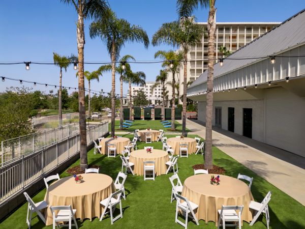 An outdoor event setup with round tables covered in beige tablecloths, white chairs, string lights, palm trees, and buildings in the background.