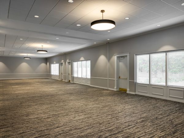 The image shows a large, empty room with carpeted flooring, multiple windows, and ceiling lights, creating a bright and open space.