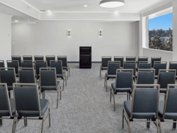 A small conference room with rows of gray chairs facing a podium at the front, illuminated by ceiling lights and natural light from a large window.