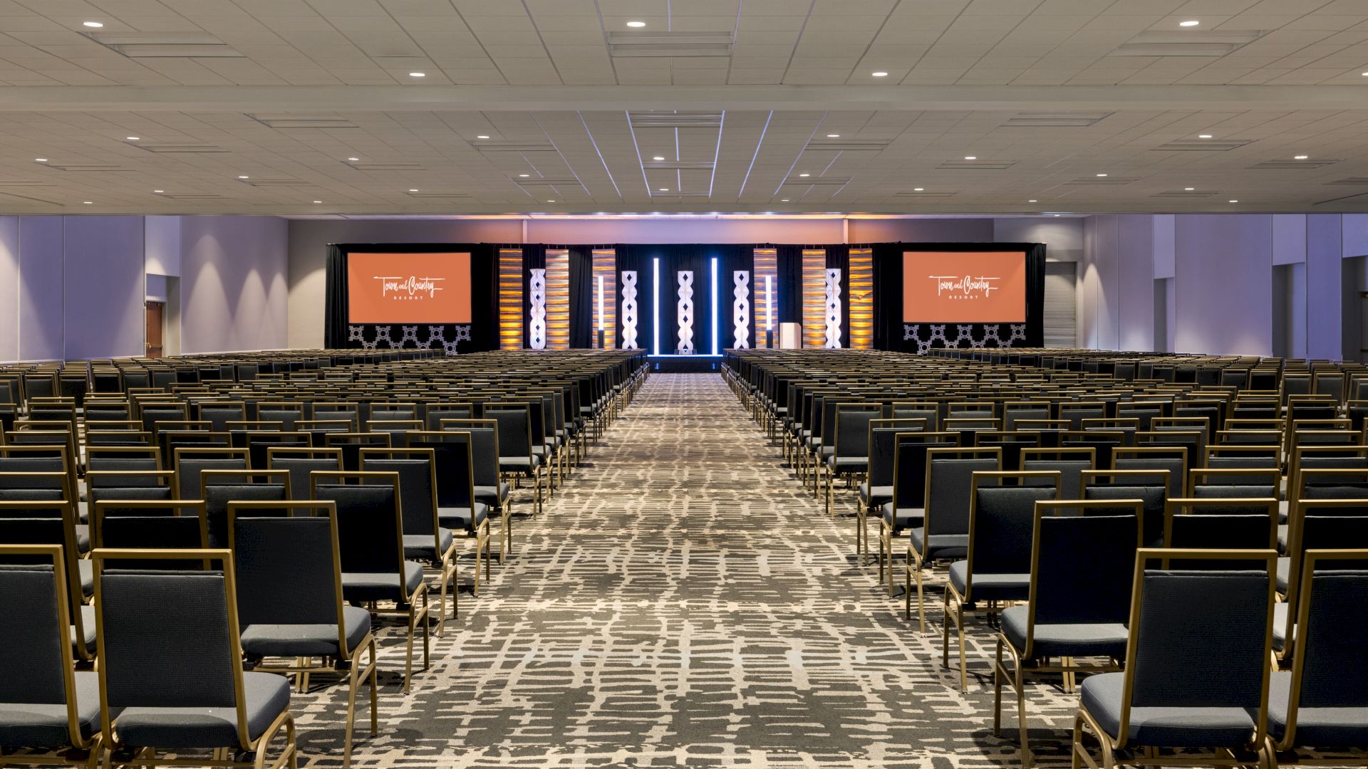A large conference room with rows of chairs facing a stage with two screens on either side, featuring a patterned carpet and overhead lighting.