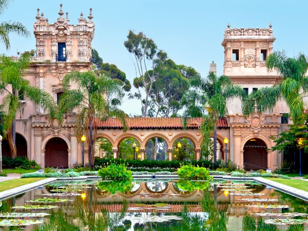 This image depicts an ornate, historic building with Mediterranean-style architecture, surrounded by palm trees and reflected in a tranquil pond.