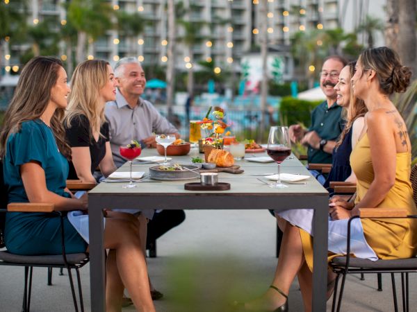 A group of six people are sitting at an outdoor dining table, enjoying food and drinks, with city lights and trees in the background.