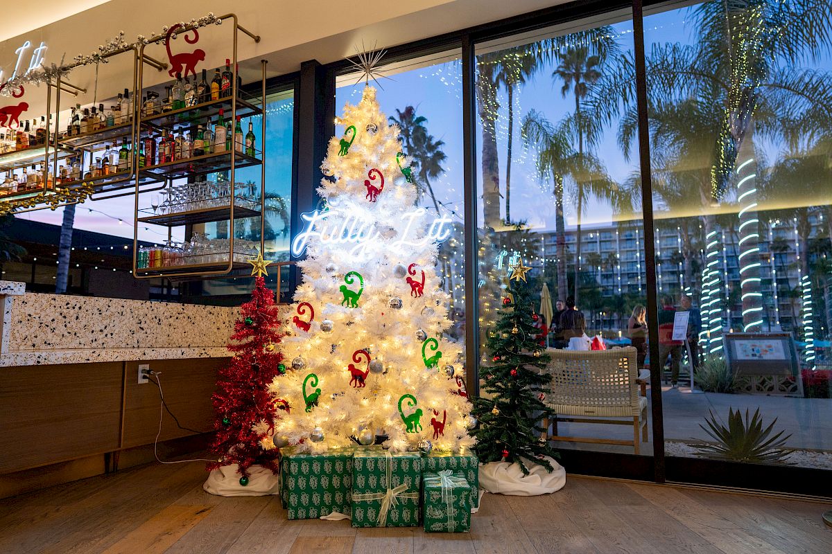 A brightly lit Christmas tree with colorful ornaments and gifts stands by a hotel bar, while holiday lights twinkle outside.