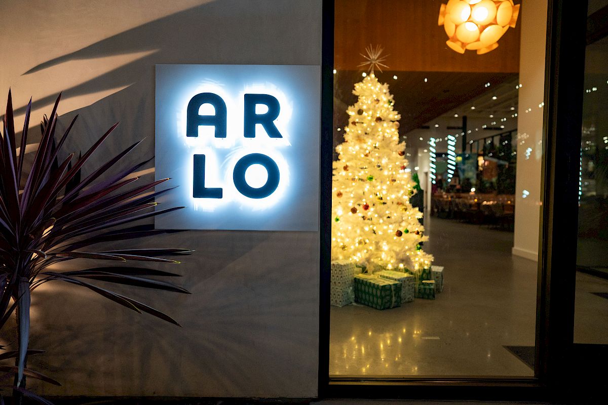 A night entrance with a glowing &ldquo;ARLO&rdquo; sign, a lit Christmas tree, and wrapped gift boxes.
