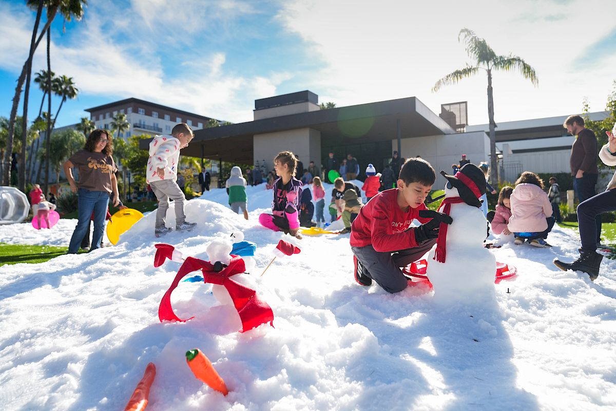 Kids play in a sunny outdoor snow area, crawling and throwing snow on foam animals while others watch and join in.