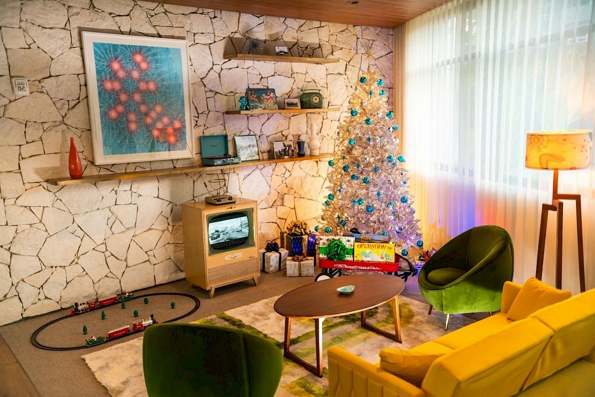 A cozy room with a decorated Christmas tree, gifts, framed art, a vintage TV, and colorful chairs.