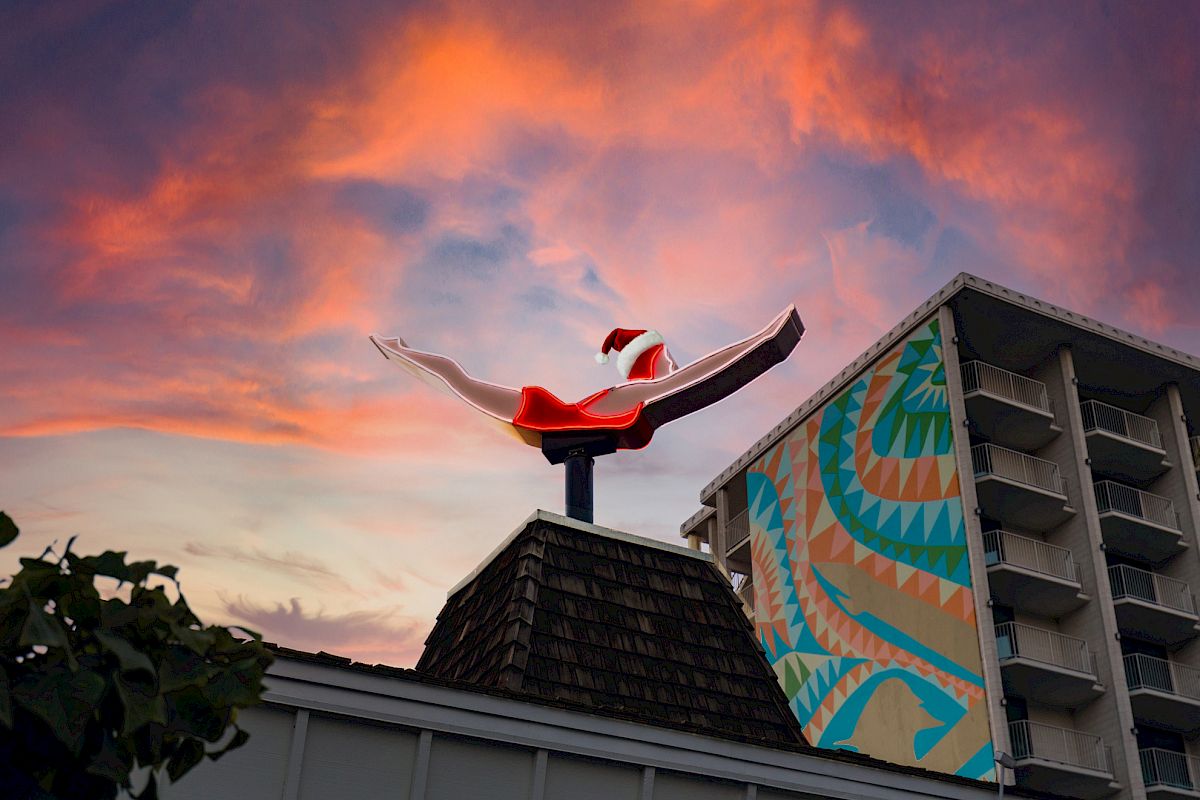 A rooftop reindeer figurine wearing a Santa hat flies through a colorful sunset beside a patterned hotel wall.
