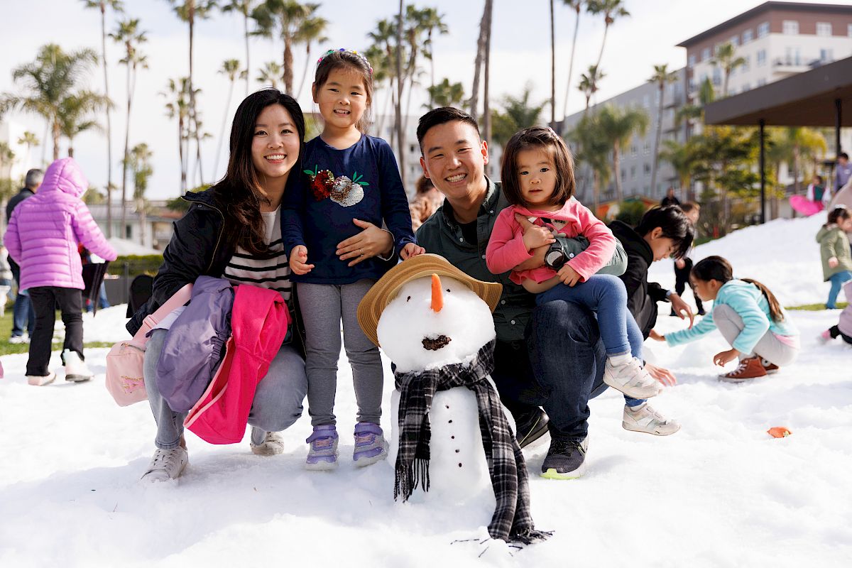 A smiling family poses in the snow with a snowman outside, while kids play in the background.