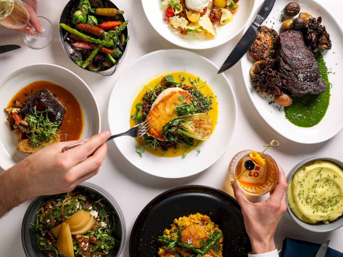 An overhead view of various gourmet dishes and hands holding a fork and drink, featuring meats, vegetables, and mashed potatoes on plates.