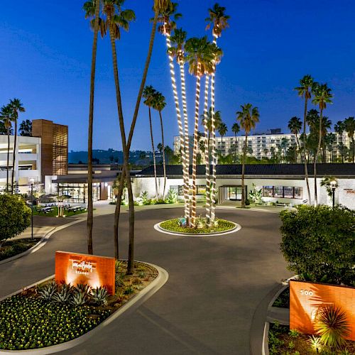 A beautifully lit hotel entrance with palm trees, a modern sculpture, lush landscaping, and a building mural at dusk, creating a serene ambiance.