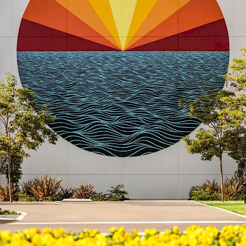 A colorful mural on a building depicts a sun setting over water with a field of yellow flowers in the foreground, flanked by trees and bushes.